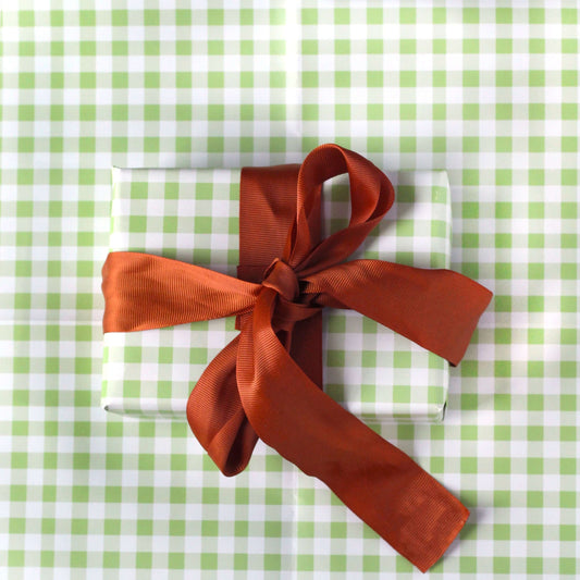 A present wrapped in green gingham wrapping paper, with an orange ribbon tied around it. The gift is sat on a sheet of the same green gingham wrapping paper.