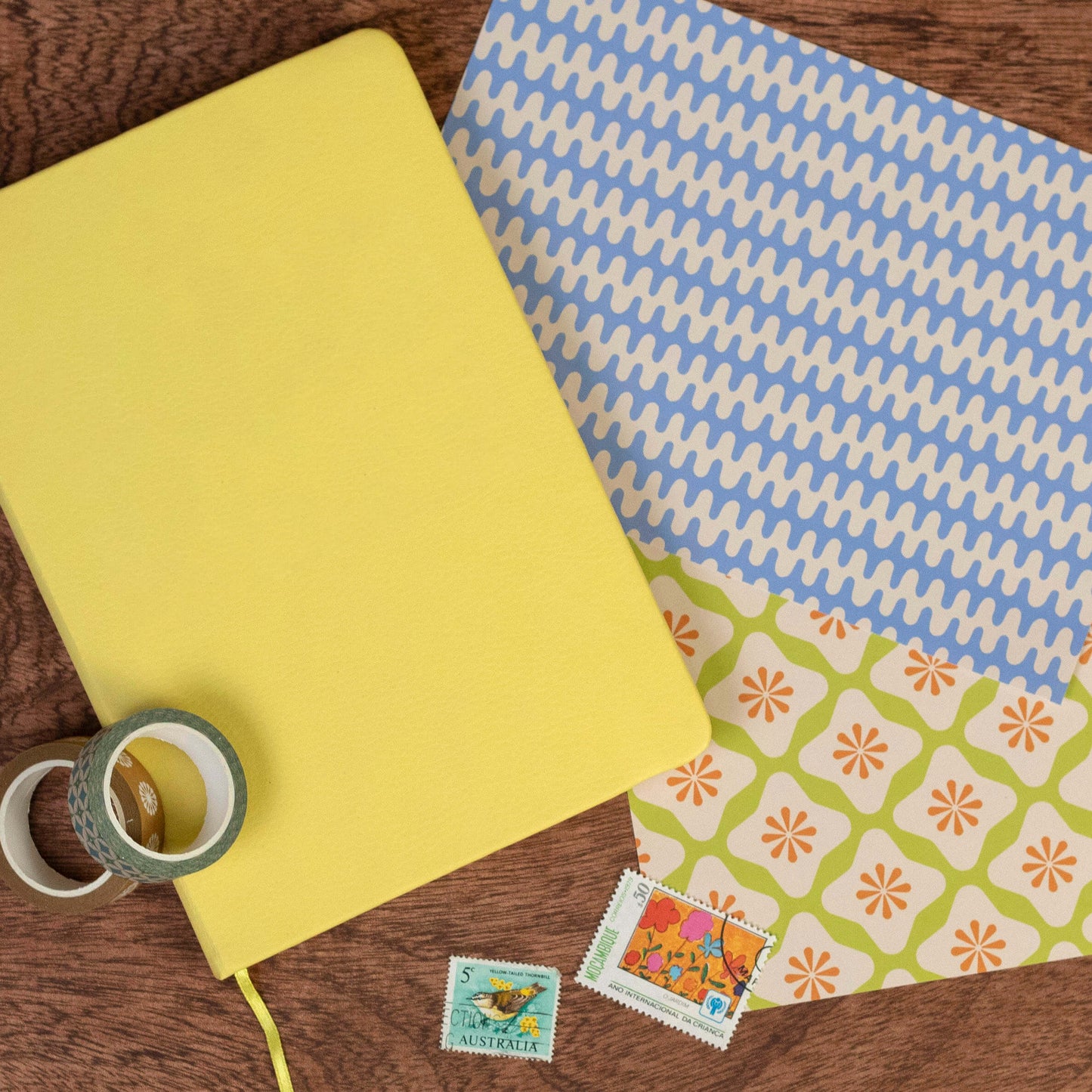 Yellow notebook, blue patterned paper, green tape, and colorful stamps on a wooden surface
