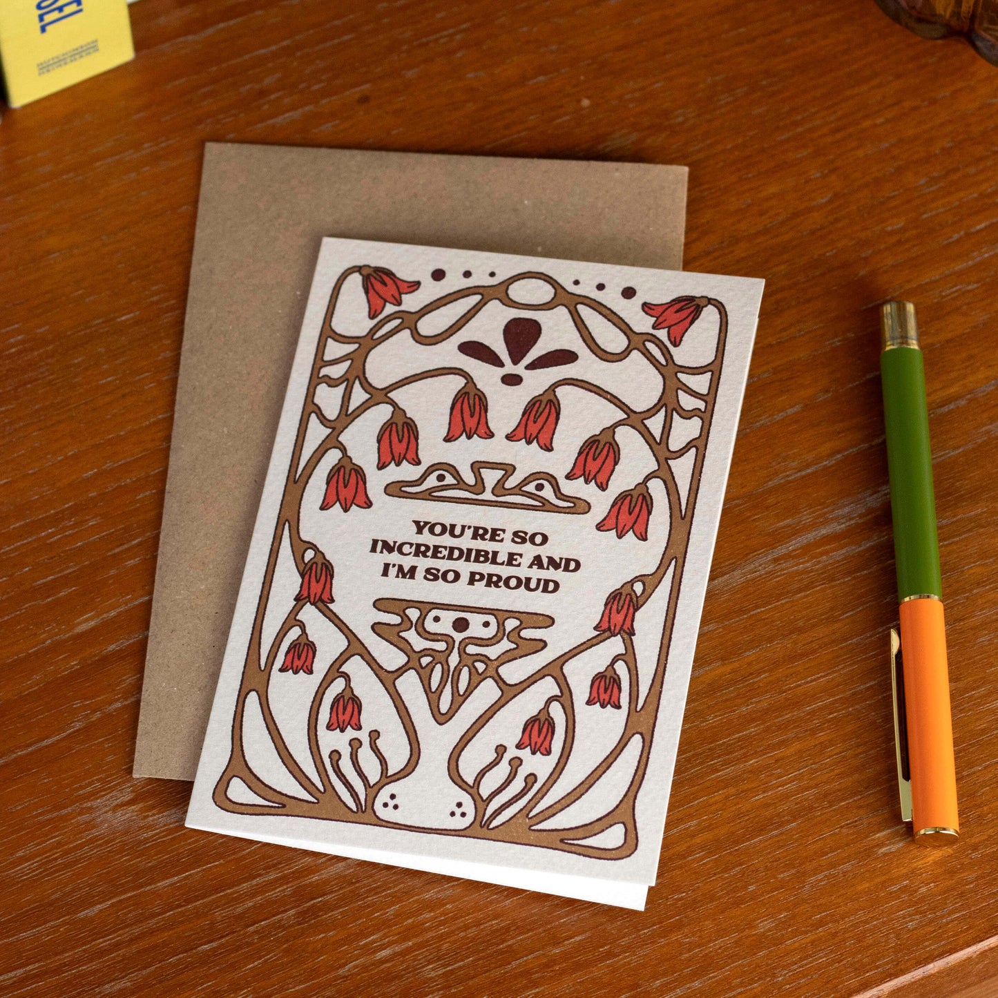 Greeting card with floral design and text saying' You're So Incredible and I'm So Proud' on a wooden surface with a pen.