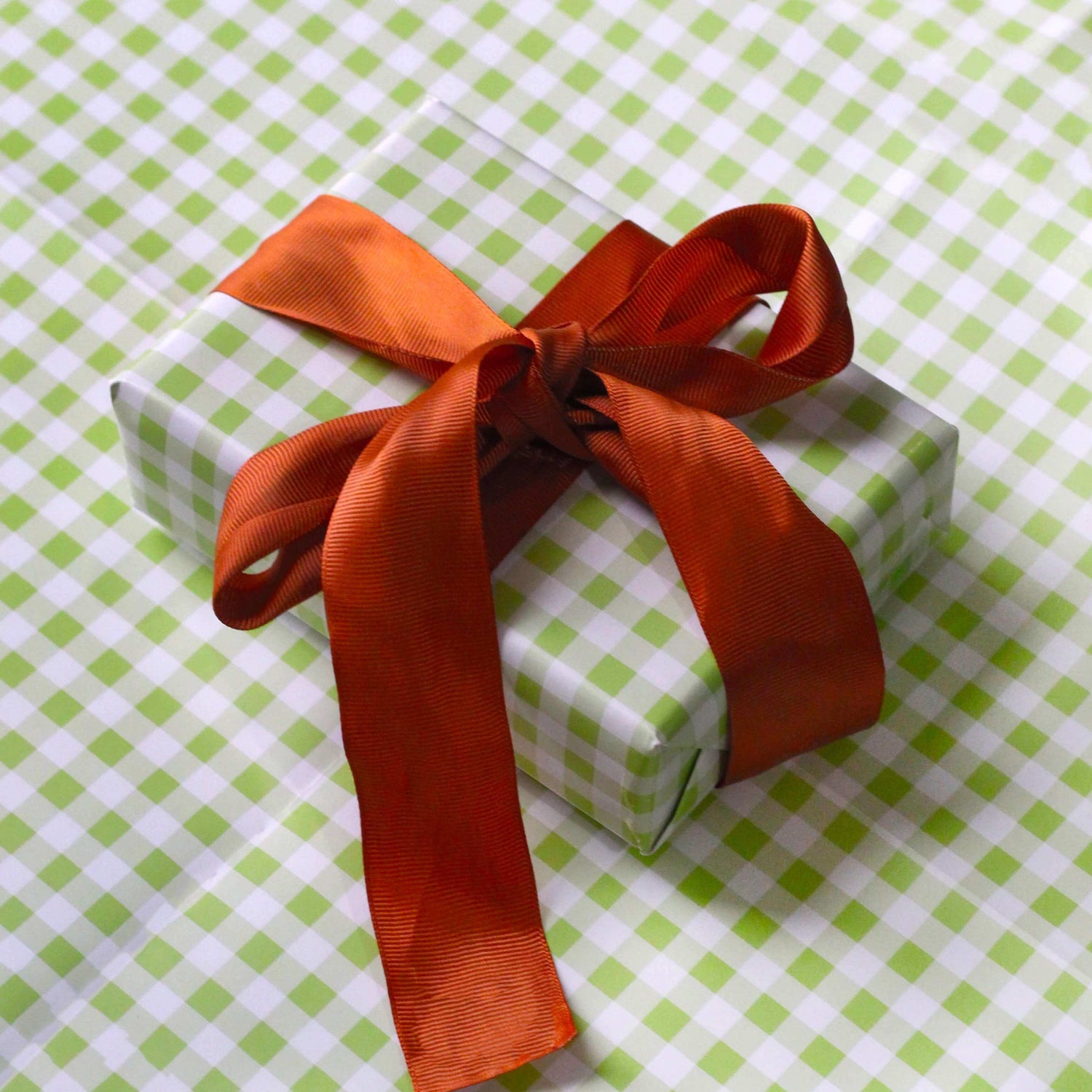 A rectangle shaped gift wrapped in green gingham wrapping paper. Behind the gift is more of the same wrapping paper, and the gift is wrapping in a large orange ribbon tied in a bow.