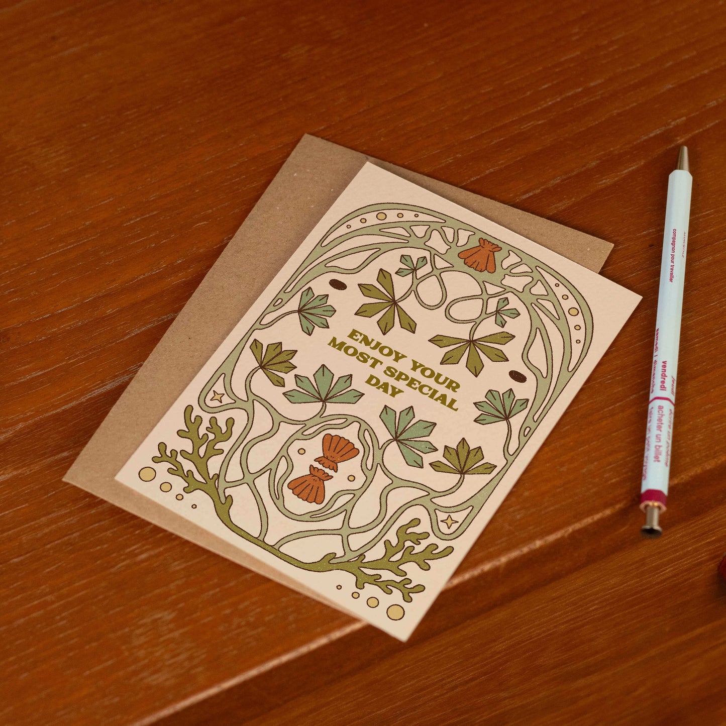 Wedding card and envelope sat on the edge of a wooden desk. There is a pale blue pen next to the card.