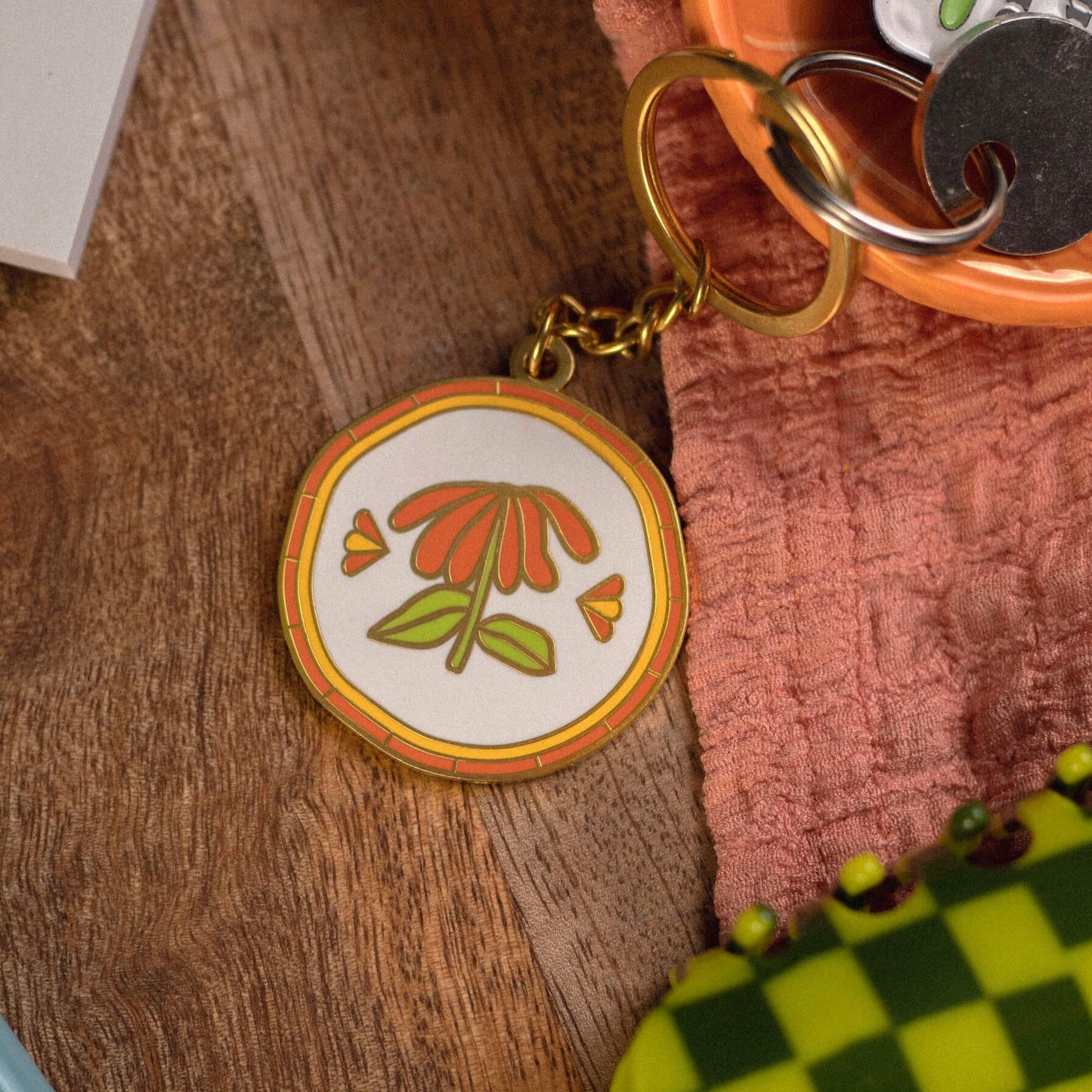 A flower keychain with gold details. It is round with an orange and yellow border, there is a single orange flower in the centre of the keychain.