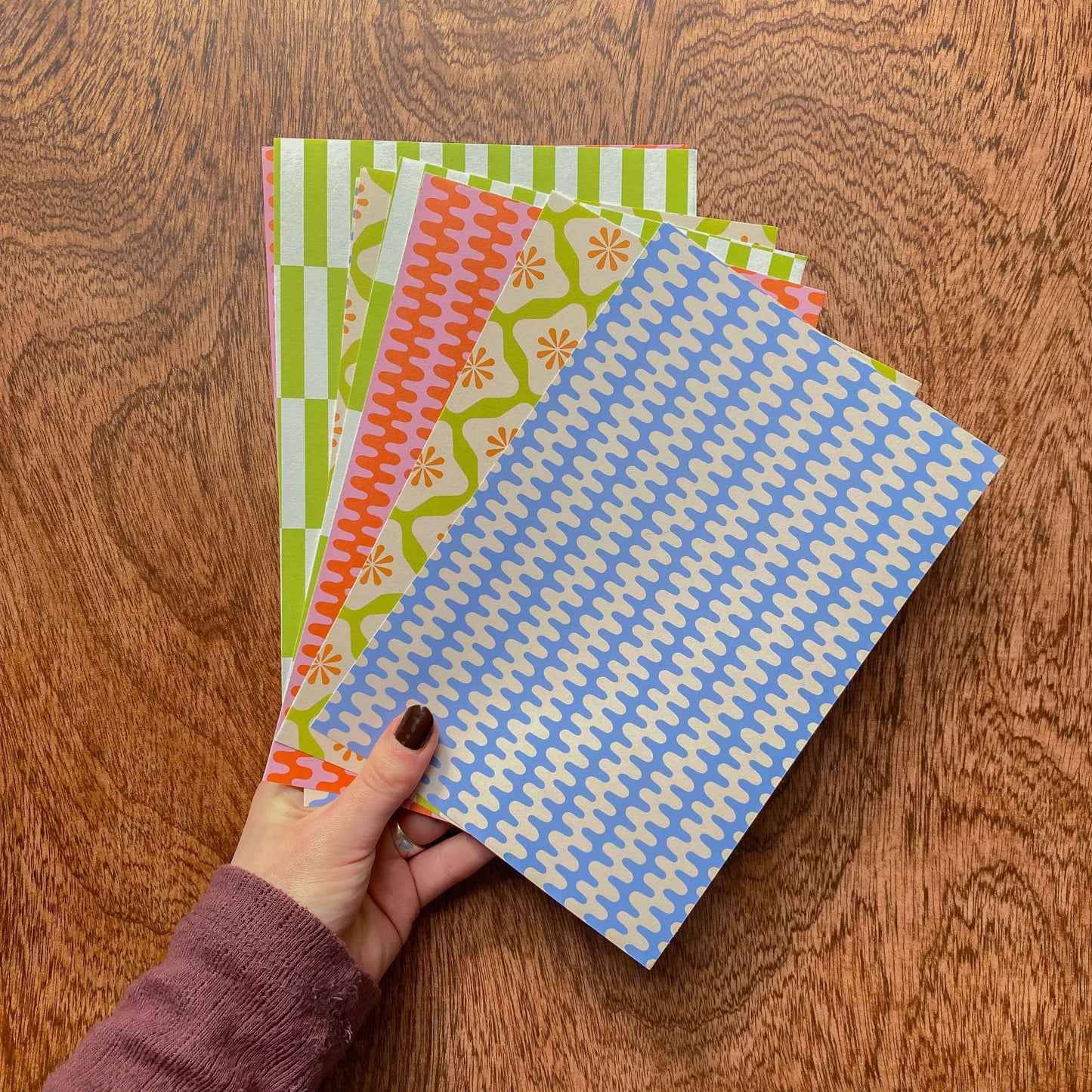 8 sheets of colourful crafting paper held by a hand in front of a wooden background