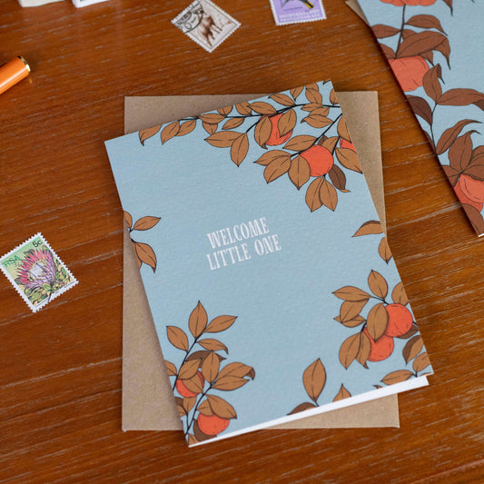 Greeting card with orange tree design  and 'Welcome Little One' text on a wooden surface surrounded by stamps.