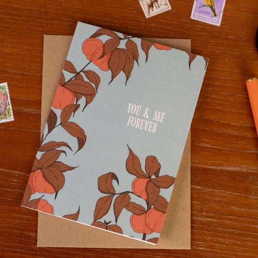 Greeting card with peach tree design and 'You & Me Forever' text on a wooden surface.